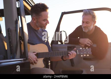 Glückliches kaukasisches, schwules Paar, das Gitarre spielt und lächelt und mit dem Auto am Meer rumhängt Stockfoto