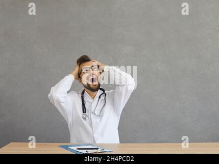 Verrückter überarbeiteter kaukasischer Arzt, der müde fühlt, schreit laut und ergreifend seinen Kopf. Stockfoto