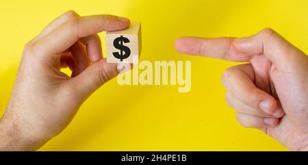 Die Hand hält einen Holzwürfel mit einem Dollarzeichen und zeigt mit einem Finger auf diesen auf einem gelben Hintergrund Stockfoto