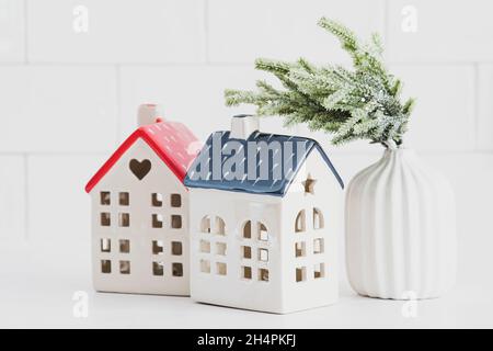 Weihnachtliche Einrichtung auf weißem Tisch. Zwei kleine Keramikhäuser und Vase mit Tannenzweig. Gemütliches Zuhause, Hygge, nordischer Stil. Stockfoto