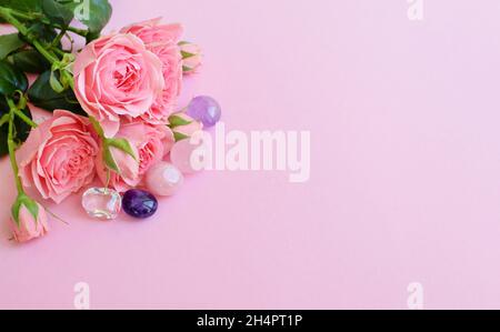 Schöne rosa Rosenblüten in voller Blüte mit grünen Blättern. Blumenstrauß auf rosa Hintergrund mit Amethysten, Rosenquarz und Bergkristall Stockfoto