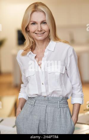 Lady Boss. Erfolgreiche reife Frau mit blondem Haar lächelt vor der Kamera, während sie drinnen steht Stockfoto
