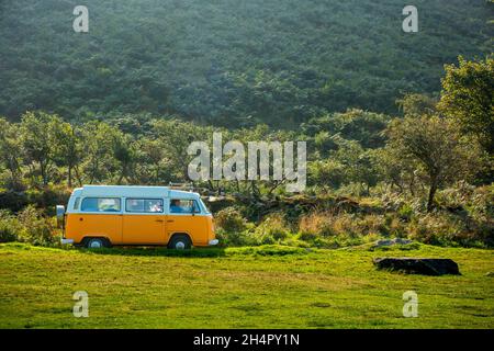 Klassischer Retro Volkswagen VW Wohnmobil fährt im Sommer durch die Landschaft mit Bäumen im Hintergrund und Gras im Vordergrund. Stockfoto