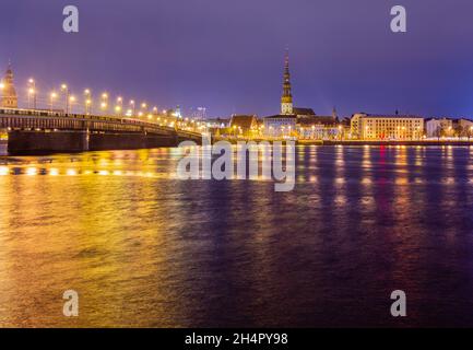 Nachtansicht des historischen Stadtzentrums und der Steinbrücke vom Ufer des Daugava Flusses mit Straßenlaternen, Riga, Lettland Stockfoto