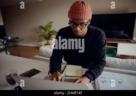 Afroamerikanischer Mann, der von zu Hause aus auf seinem Laptop arbeitet. Afroamerikanische Planungswoche für Männer mit Papier und Stift Stockfoto