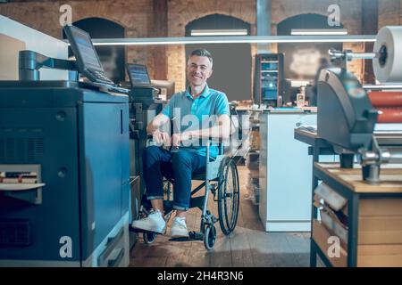 Mann im Rollstuhl, der die Kamera in der Nähe von Bürogeräten ansieht Stockfoto