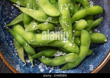 Biologischer gedämpfter japanischer Edamame, eine junge Sojabohne, die reich an Nährstoffen ist, serviert in einer blauen Schale und mit Meersalzflocken bestreut. Gemüse Backgr Stockfoto