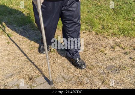 Behinderter älterer Mann mit Gehstock, der einen harten Schritt auf einem bewachsenen Gehweg mit Gras macht Stockfoto