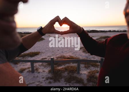 Glücklich kaukasisch Gay Männlich Paar bilden Herzform mit Händen am Strand bei Sonnenuntergang Stockfoto