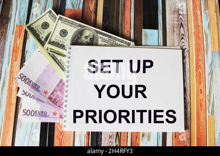 Dollar- und Euro-Banknoten liegen auf einem gestreiften Hintergrund. Text auf einem weißen Blatt Notizblock.Setzen Sie Ihre Prioritäten. Stockfoto