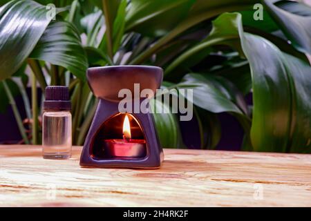 Eine aromatische Lampe mit einer Kerze im Inneren steht auf einer hölzernen Tischplatte vor einem Hintergrund aus grünen Blättern. Stockfoto