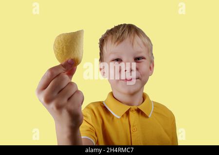 Ein glücklicher Junge hält einen Kartoffelchip in der Hand auf einem gelben Hintergrund. Stockfoto