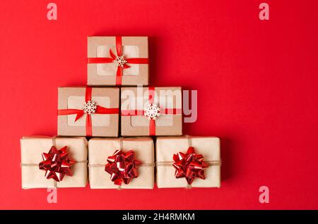 Weihnachtsgeschenke werden in Form eines Baumes auf einem roten Hintergrund angelegt. Geschenke in kraftkoroboki und Kraftpapier mit Bögen Stockfoto