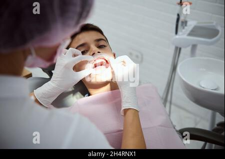 Überkopfansicht eines pädiatrischen Zahnarztes, der vor der oralen Behandlung einen Wangenaufroller auf einen Kinderpatienten legt. Schuljunge beim Zahnarzttermin. Den Stockfoto