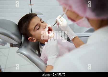 Selbstbewusster Kinderjunge, der auf dem Zahnarztstuhl in der weißen modernen Zahnklinik sitzt, während eine nicht erkennbare weibliche Zahnärztin medizinische Manipulationen und Stockfoto