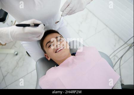 Liebenswert tapfere gut aussehende vorpubertär Junge im Zahnarztstuhl lächelt toothy Lächeln Blick auf Kamera und wartet auf zahnärztliche Untersuchung und Behandlung in Stockfoto