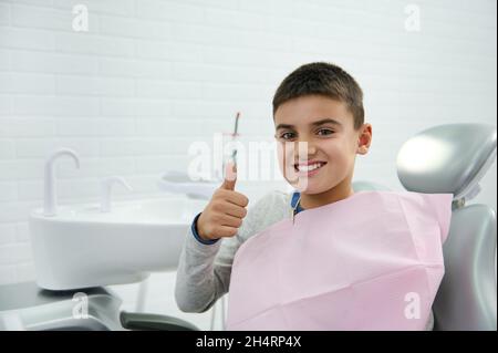 Fröhlich selbstbewusst tapfer gutaussehender Schuljunge, Kind im Grundschulalter zeigt Daumen nach oben und lächelt mit einem schönen toothy Lächeln auf die Kamera, während sie sitzt Stockfoto