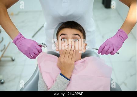 Draufsicht auf Angst Kind Junge seinen Mund vor Angst mit der Hand, sitzt auf dem Zahnarztstuhl auf dem Hintergrund der Hände in rosa OP-Handschuhe Hol Stockfoto