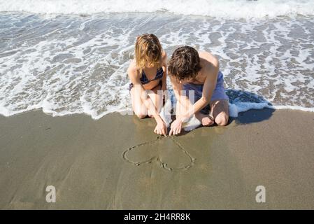 Ein junges verliebtes Paar, eine Frau und ein Mann, ziehen ein Herz auf den Sand an der Küste. Frohe Feiertage, erste Liebe, glückliche Beziehungen, Sommerurlaub, Stockfoto