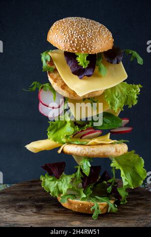 Schwebender Burger. Fliegende Nahrung. Hamburger-Brötchen, doppeltes Hühnerschnitzel mit Käse, Salat, Rucola und Rettich. Fleisch, Gemüse und Brot. Zutaten auf schwarzem Hintergrund. Stockfoto