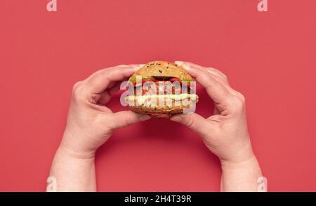 Draufsicht mit Frauenhänden, die einen kleinen veganen Burger halten. Gesundes Essen, ein hausgemachter Cheeseburger mit Roggenbrotbrötchen und Sojafleischpastete. Stockfoto