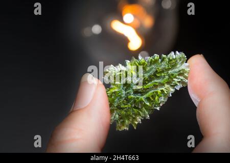 Seltener grüner Moldavit-Edelstein in menschlichen Fingern mit Glühlampe auf schwarzem Hintergrund. Nahaufnahme von schönem Edelstein aus faltenem kometenhaften Glas. Stockfoto