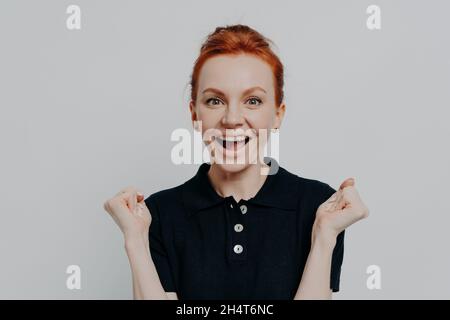 Glückliches junges Ingwer-Mädchen, das von tollen Neuigkeiten begeistert ist und mit geballten Fäusten die Hände hebt Stockfoto