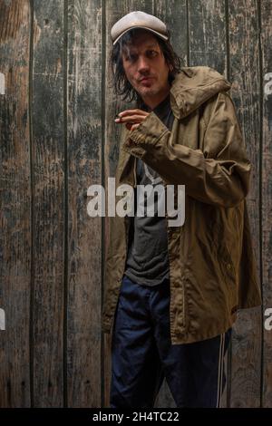 Junkie-Mann mit Mütze grüner Anorak und blauer Jogginghose in der Nähe einer alten hölzernen Wand Stockfoto