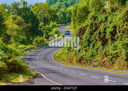 Ein Auto fährt auf einer Straße. Eine schmale, kurvenreiche Straße in Schottland entlang des Loch Ness. Bäume und Sträucher neben der Straße im Sommer bei Sonnenschein. Verkehrszeichen und Stockfoto