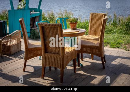 Ein Holztisch und vier Korbstühle - Gartenmöbel stehen auf einer hellen Holzterrasse am Flussufer. Sonniger Sommertag. Stockfoto