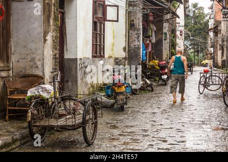 Ein Mann geht in Xingping, China, eine schmale Straße entlang, die mit Fahrrädern und Motorrädern gesäumt ist. Stockfoto