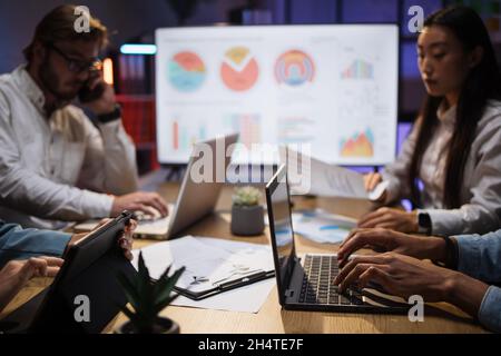 Nahaufnahme von Mitarbeitern multiethnischer Unternehmen, die spät im Büro sitzen und moderne Gadgets verwenden. Konzentrieren Sie sich auf den nicht erkennbaren afroamerikanischen Typ, der auf einem modernen Laptop tippt Stockfoto