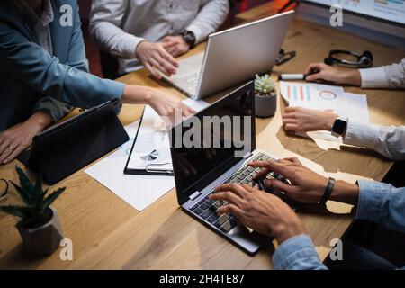 Nahaufnahme aus dem oberen Blickwinkel von Mitarbeitern aus mehreren ethnischen Unternehmen, die spät im Büro sitzen und moderne Gadgets verwenden. Männer und Frauen analysieren den Finanzbericht während der Sitzung. Stockfoto