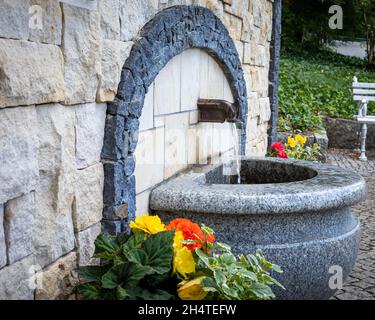Trinkbrunnen umgeben von Blumen. Steinbecken, Wasser läuft aus dem Wasserhahn. Ladek Zdrój Mineral Water Kurort, Polen. Stockfoto