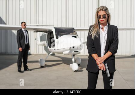 Schöne professionelle weibliche Spionageagent Bodyguard mit Pistole Sicherung Flugzeug auf dem privaten Flughafen. Frau der Sicherheitspolizei in zivilem schwarzen Anzug. Schließen Sie s Stockfoto