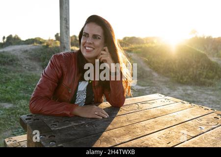 Hübsche lächelnde Frau, die bei Sonnenuntergang auf einem Tisch in einem Park sitzt und eine rote Jacke trägt Stockfoto