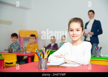 Nettes präteen Mädchen studieren im Klassenzimmer auf Hintergrund mit Klassenkameraden und Lehrer Stockfoto
