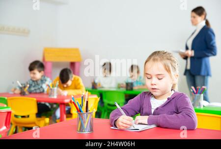 Nettes präteen Mädchen studieren im Klassenzimmer auf Hintergrund mit Klassenkameraden und Lehrer Stockfoto
