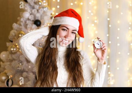 Schöne Frau in santa Hut hält einen Wecker und lächelt niedlich vor dem Hintergrund der festlichen weihnachtsbaum mit Lichtern von Girlande. Kaukasisches Mädchen in Erwartung der Wunder und Geschenke des neuen Jahres. Stockfoto