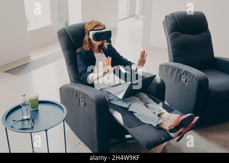 Junge moderne Geschäftsfrau in VR-Brille sitzt mit geöffnetem Laptop auf einem Sessel zu Hause Stockfoto
