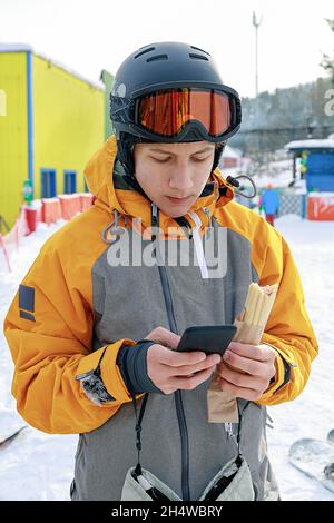 Junge Snowboarderin mit einem Smartphone. Ein Kerl in einem hellen Sportoverall und einem Schutzhelm auf der Skipiste. Junger Mann schreibt eine Textnachricht. Mobi Stockfoto