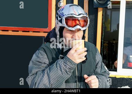 Porträt eines Snowboarders mit einer Tasse heißen Kaffees aus Papier. Ein Mann aus der kaukasischen Volkszugehörigkeit mittleren Alters in einem Schutzhelm, der sich auf einer Skipiste ausruht und trinkt Stockfoto