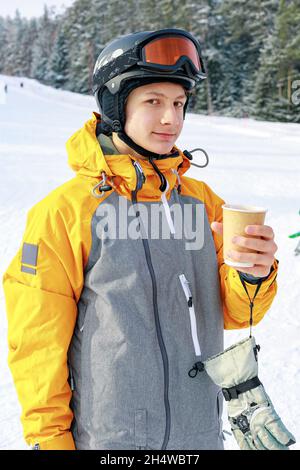 Der Snowboarder trinkt heißen Kaffee. Ein junger Mann aus dem Kaukasus hält einen Papierbecher mit einem heißen Getränk in der Hand. Hintergrund der Skipiste. Porträt eines AT Stockfoto