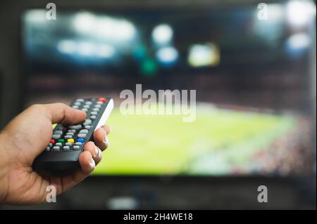 Junger Mann mit Fernbedienung und Fußballprogramm. Hände, die auf den fernseher zeigen und ihn ein- oder ausschalten, wählen Sie die Senderüberwachung aus Stockfoto