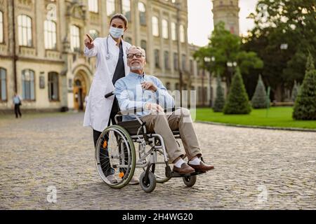 In voller Länge Aufnahme einer fürsorglichen jungen Krankenschwester in Schutzmaske, die sich um einen behinderten älteren Patienten im Rollstuhl im Freien kümmert Stockfoto