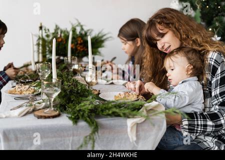 Am Weihnachtstag frühstücken Mama und die Kinder morgens an einem festlich geschmückten Tisch im hellen Wohnzimmer. Die Familie isst am Tisch Stockfoto