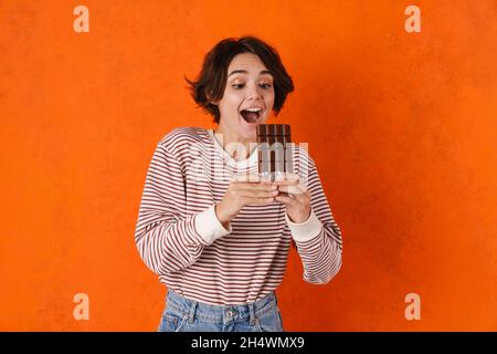 Junge Brünette Frau lächelt beim Essen Schokolade isoliert über orange Wand Stockfoto
