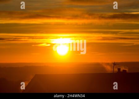 Der Dampf aus zentralen Heizgaskesseln wird durch den Herbstaufgang in Northowram, Calderdale, West Yorkshire, Großbritannien, wieder erleuchtet. Der wässrige gelbe Schein des Morgens reflektiert sich auf der Unterseite der Wolken. Stockfoto