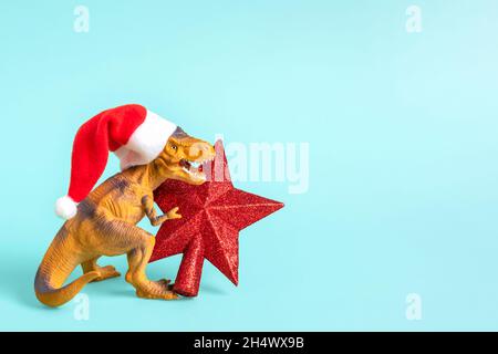 Dinosaurier Rex in rot Weihnachtsmann Hut hält Stern in den Pfoten auf blauem Hintergrund Silvester oder Heiligabend Art Weihnachtskarte Kreative Idee für Merry Stockfoto