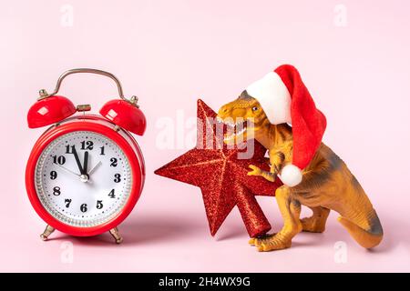 Dinosaurier Rex in rotem Weihnachtsmann Hut und Wecker auf rosa Hintergrund Silvester oder Heiligabend Art Weihnachtskarte Kreative Idee für Merry Xmas Co Stockfoto